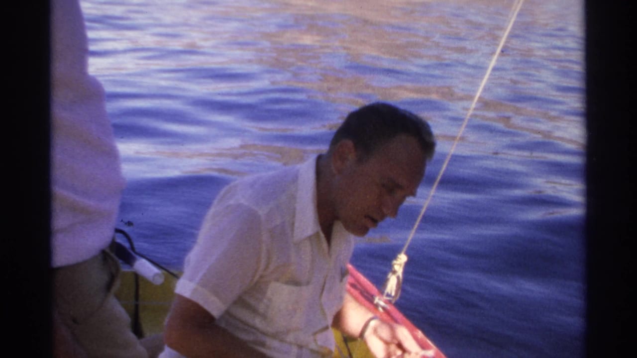 Man in a white shirt seated in a small boat on blue water, handling fishing gear.