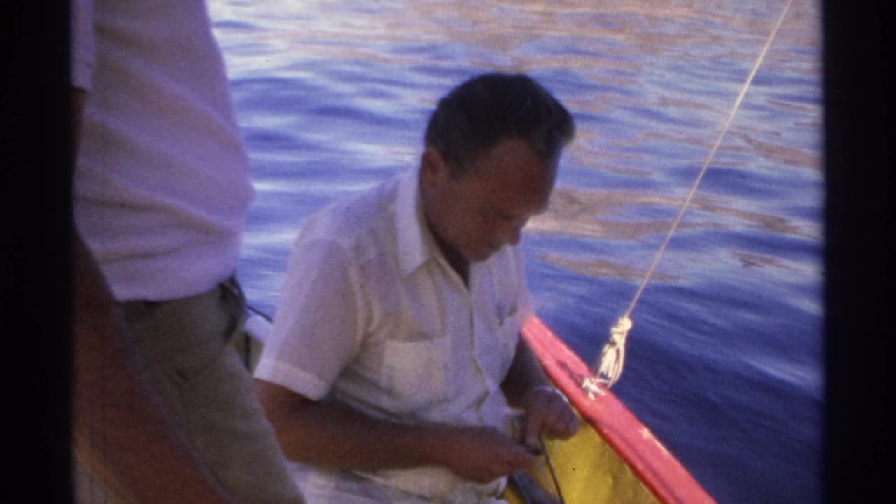1975 Guaymas frame showing a seated man concentrating on fishing gear in a small boat, with rippling blue water behind him.