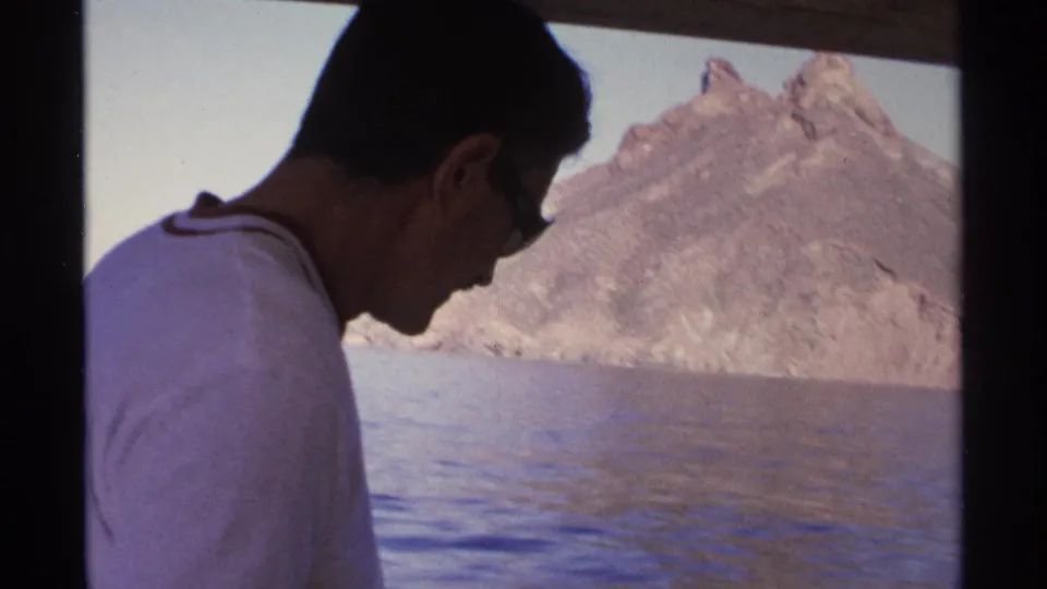 1975 Guaymas home-movie frame of a man in sunglasses seen in profile on a boat, with calm water and rocky shoreline beyond.