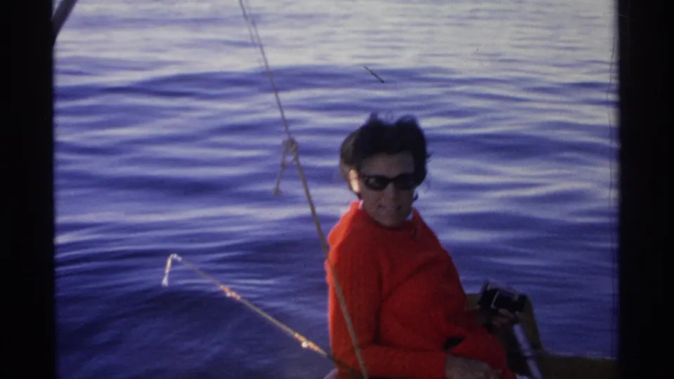 In 1975 Guaymas, Mexico footage, a woman in a red sweater and dark sunglasses sits on a boat with rippling blue water behind her.