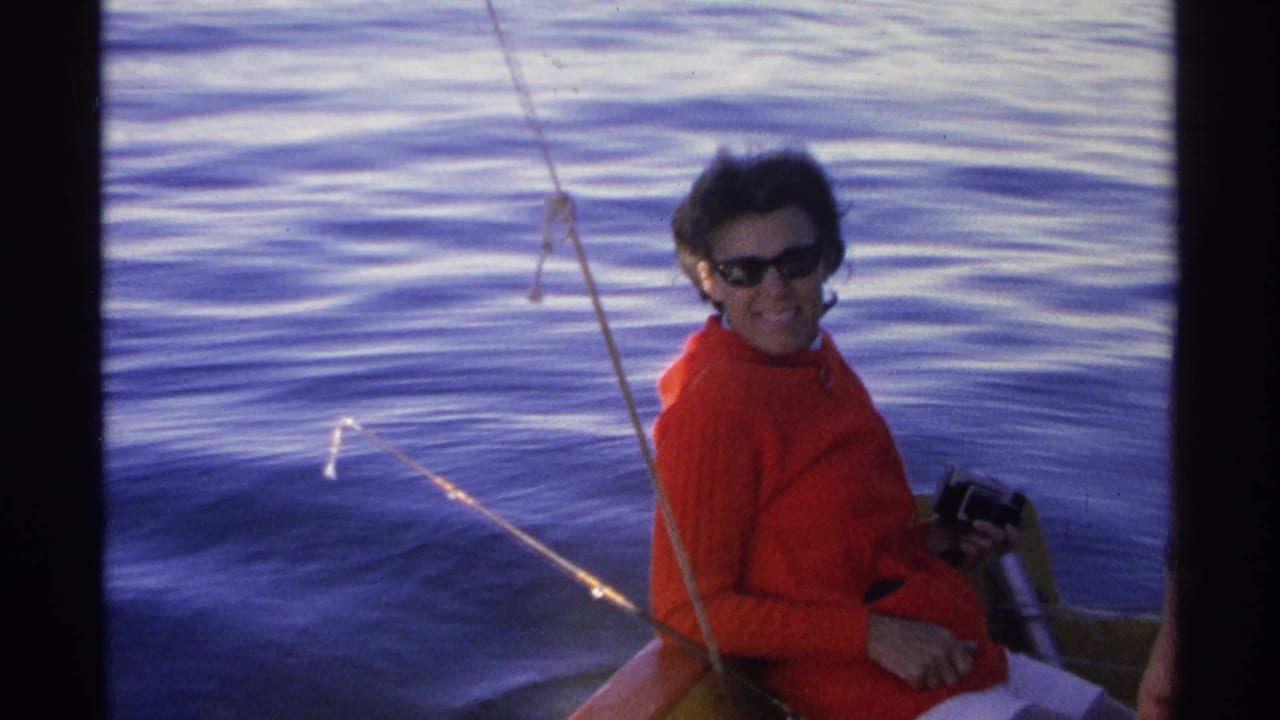 Woman in red sweater smiles from a boat while holding a camera near blue water.