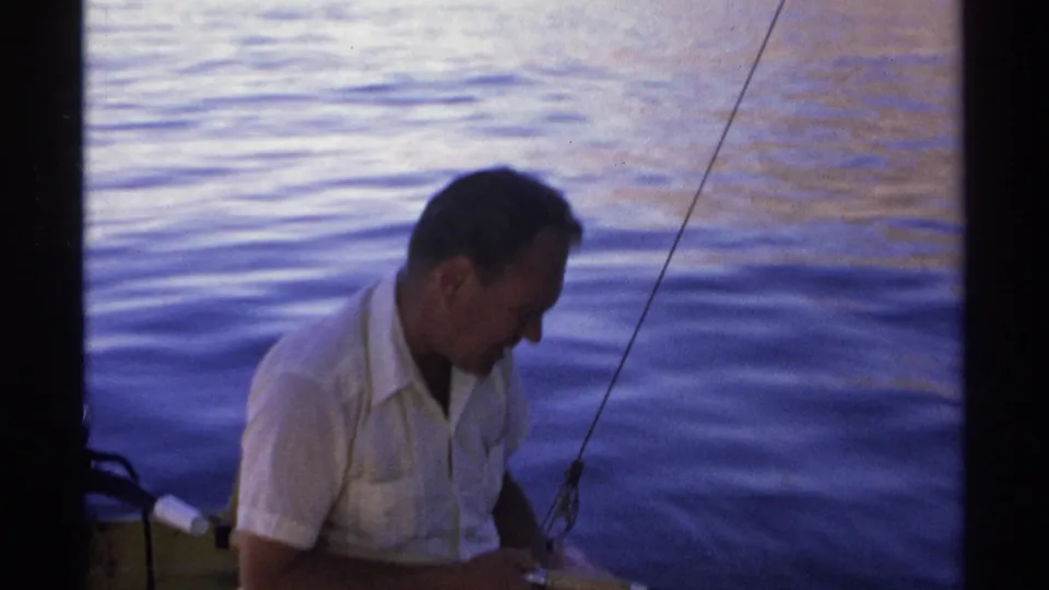 A man in a white short-sleeved shirt focuses on a fishing line from the small boat, with rippling blue water in the background.