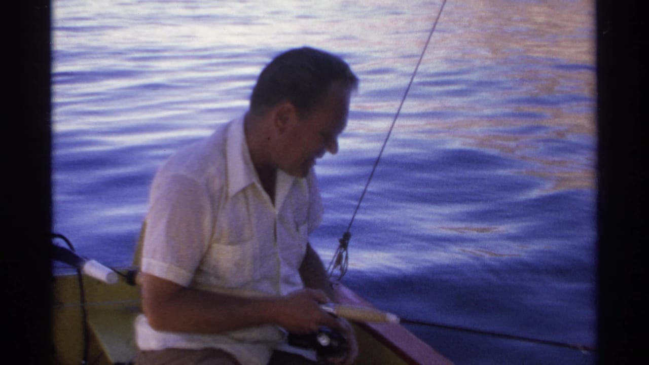 Man in a white short-sleeved shirt sits in a small boat holding a fishing rod over rippling water.