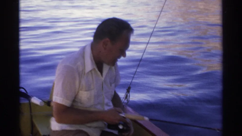 In 1975 Guaymas home-movie footage, a seated man in a white shirt handles a fishing rod beside calm blue water.