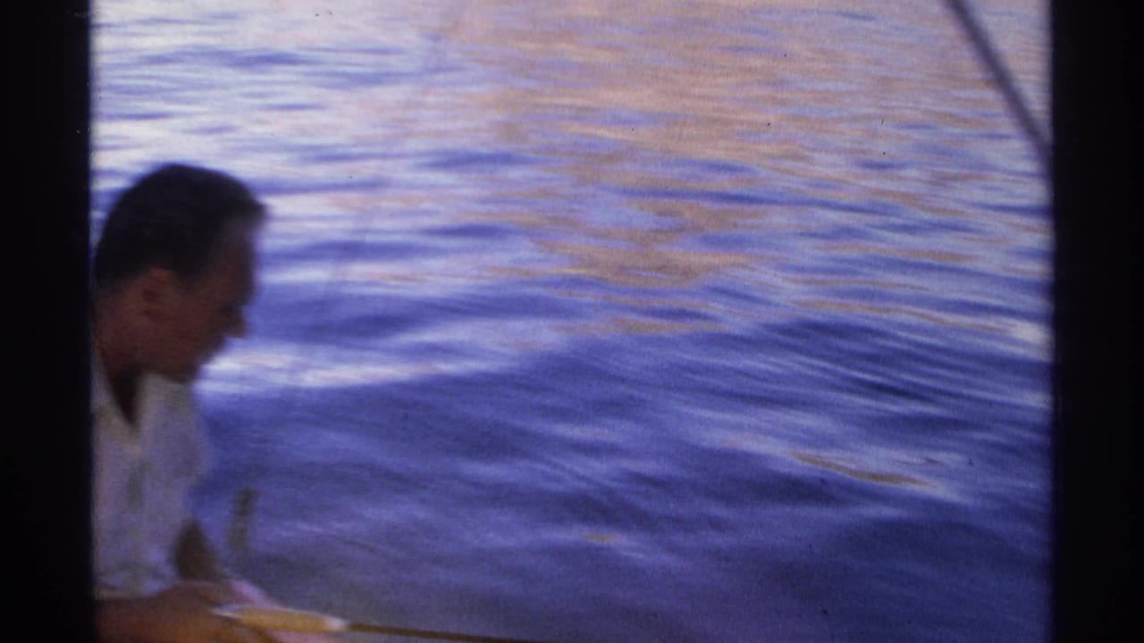 Man in profile sits low in a boat near dark rippling water with a fishing rod partly visible.