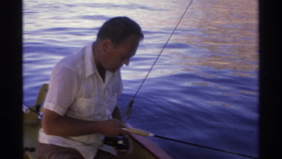 The fisherman sits near the boat edge in Guaymas, holding the reel while his rod angles across calm 1975 8mm footage.