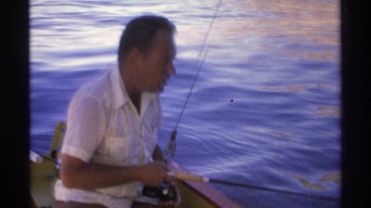 Man in a white short-sleeved shirt holds a fishing rod on a small boat beside calm blue water.