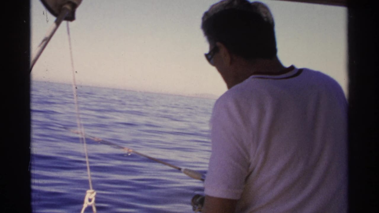 Man seen from behind fishes over calm ocean water with boat rigging in the foreground.