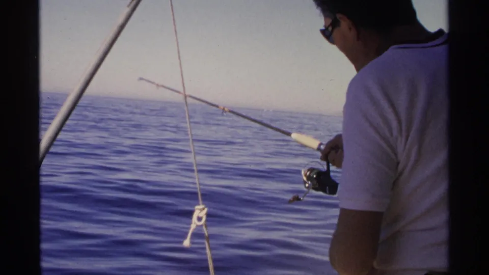 The 1975 home-movie frame shows a fisherman aboard a boat in Guaymas, Mexico, holding a rod over calm blue water.