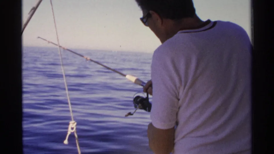 A man in a white shirt and sunglasses reels a fishing line from a boat on calm water in Guaymas, Mexico, in 1975.