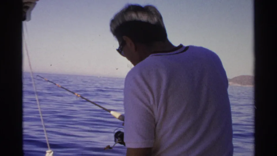 From behind, the man continues fishing from the boat, with calm water and a low coast visible in the distance.
