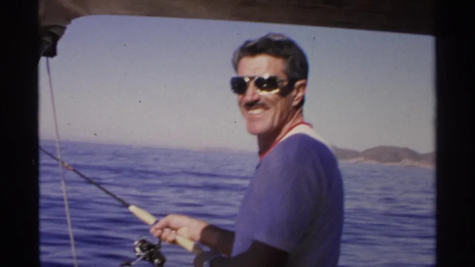 In Guaymas, Mexico, 1975, a man in aviator sunglasses smiles toward the camera while fishing from a boat on blue water.