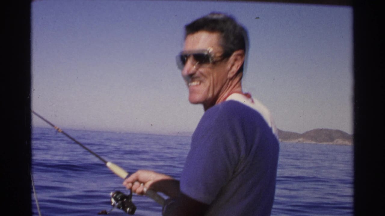 Man wearing sunglasses turns with a fishing rod against open water and hills.