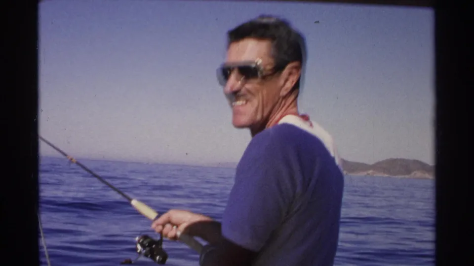 The man continues fishing from a boat in Guaymas, Mexico, with blue water, distant hills, and clear sky behind him.