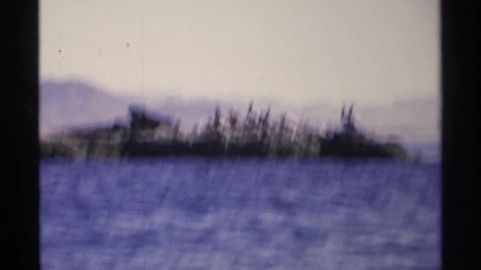 A soft-focus 1975 Guaymas coastal view shows blue water in front of a low, dark shoreline with hazy hills beyond.