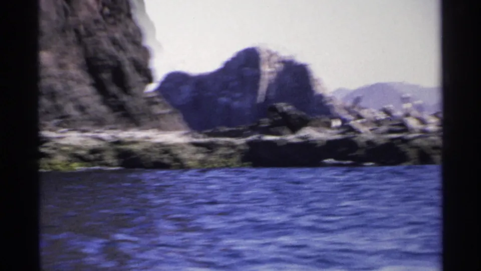 1975 Guaymas 8mm frame showing a closer rocky shoreline with pelicans, rippled blue water, and soft mountain forms in the distance.