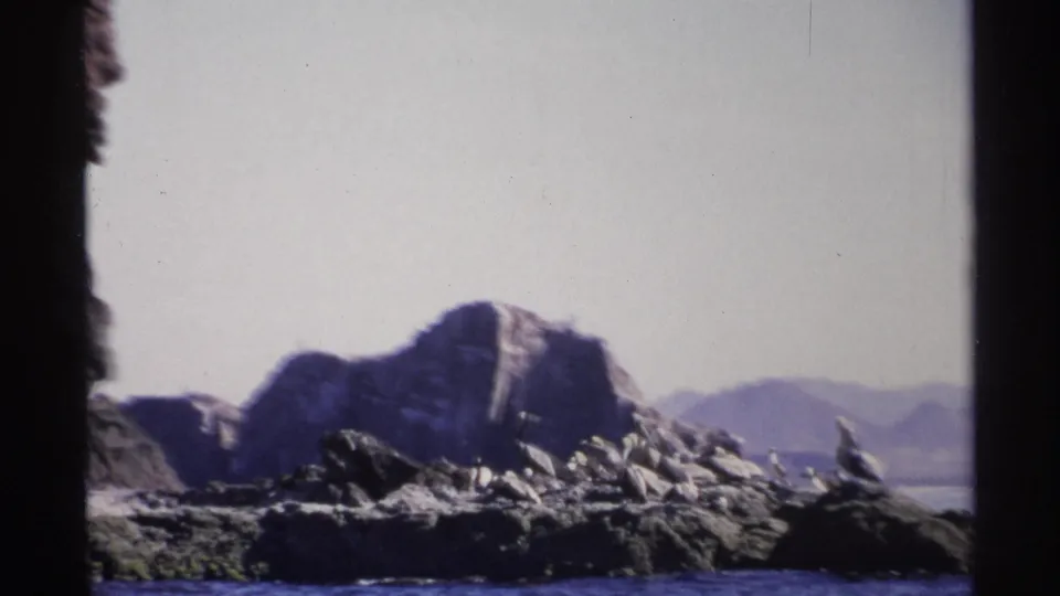 1975 Guaymas home-movie frame showing rocky shoreline and a low mountain across blue water under a pale sky.