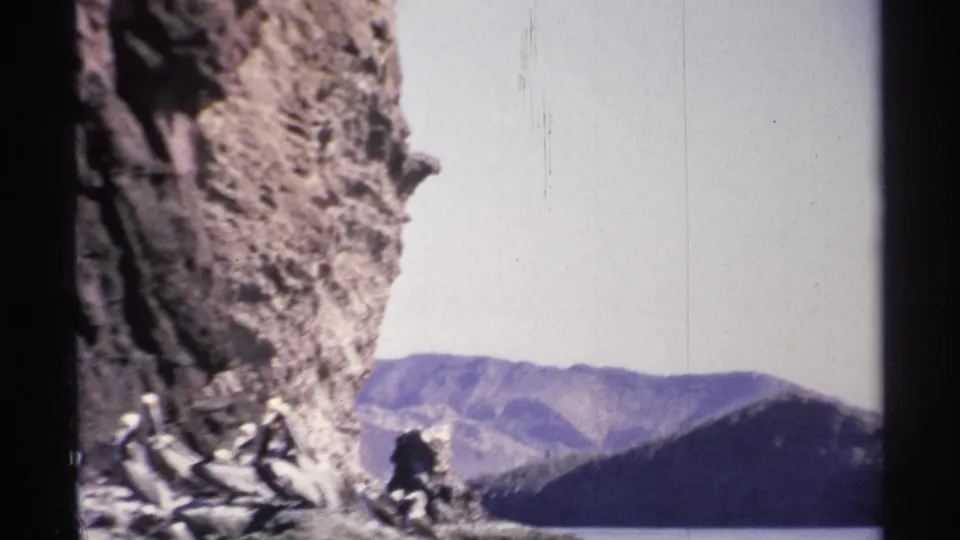 Final 1975 Guaymas frame showing seabirds at the base of a large rock formation, with blue-gray mountains in the background.