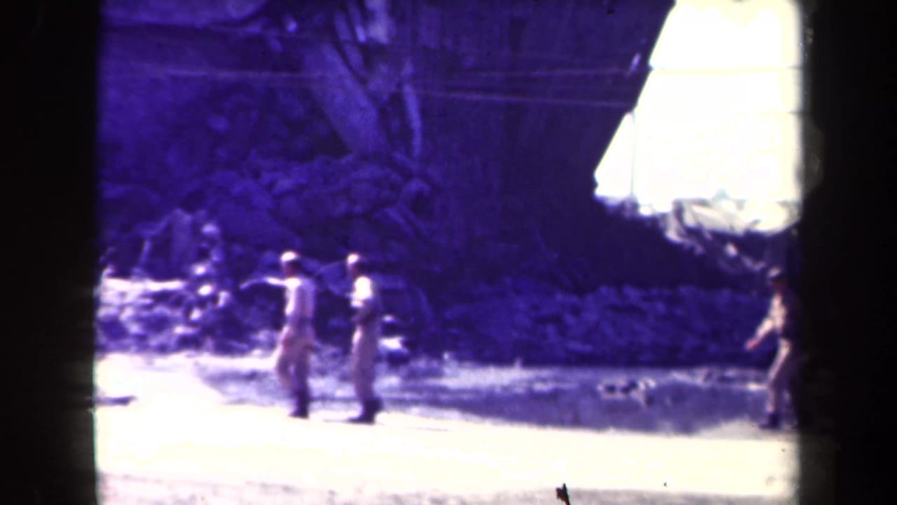 Uniformed men walk along a pale road beside a large ruined building.