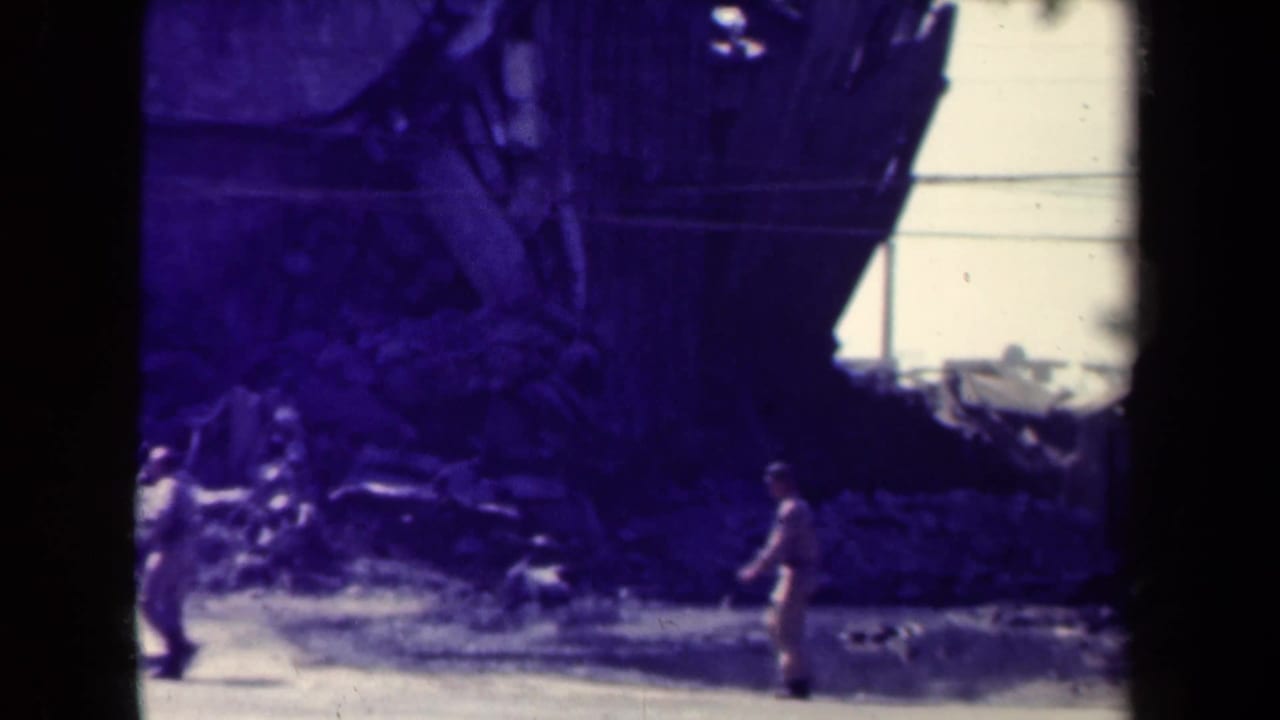 A uniformed figure in wartime Manila appears near rubble and a damaged building, with utility lines visible in the background.