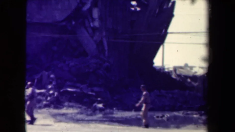 A uniformed figure in wartime Manila appears near rubble and a damaged building, with utility lines visible in the background.