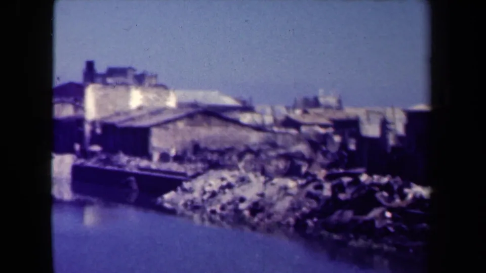 1944 Manila 8mm frame with sloped-roof buildings, a rough riverbank, and calm water in the foreground.