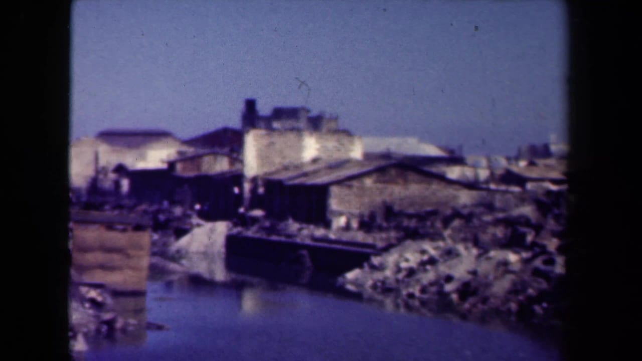 Damaged waterfront buildings and rubble beside still water, framed by dark 8mm film borders.