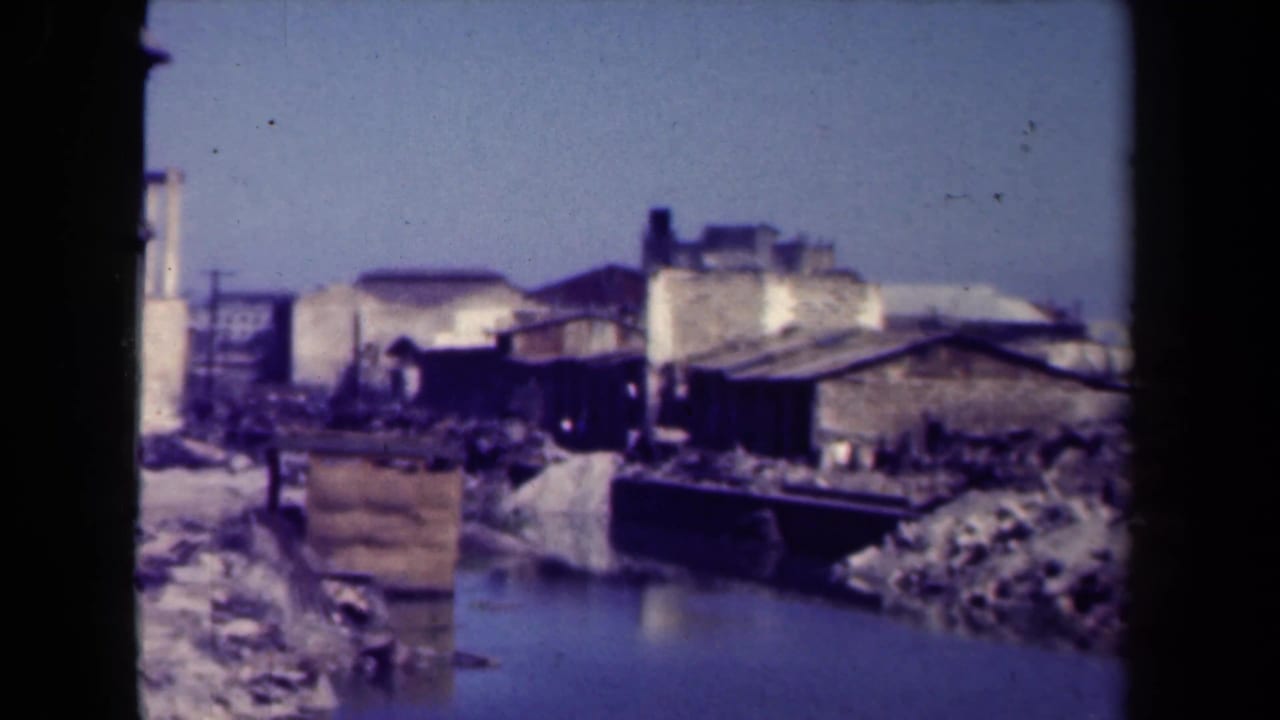 Similar view of damaged buildings and debris along a waterway with heavy film vignetting.