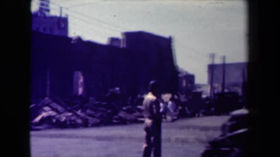 1944 Manila street view with a standing figure amid rubble, damaged buildings, poles, and a dark vehicle shape.