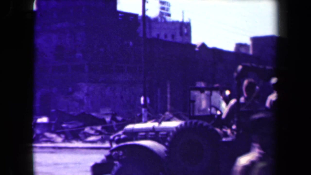 Military jeep passing a damaged city street with pedestrians and rubble in the background.