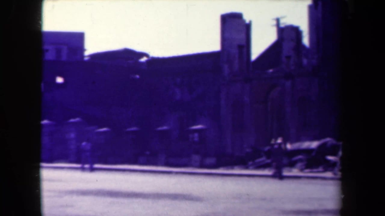 Street view of weathered stone buildings and a dark arch edge with a small figure near the roadway.
