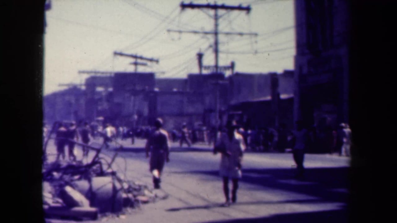 Pedestrians walking along a street with rubble, utility poles, and overhead wires.