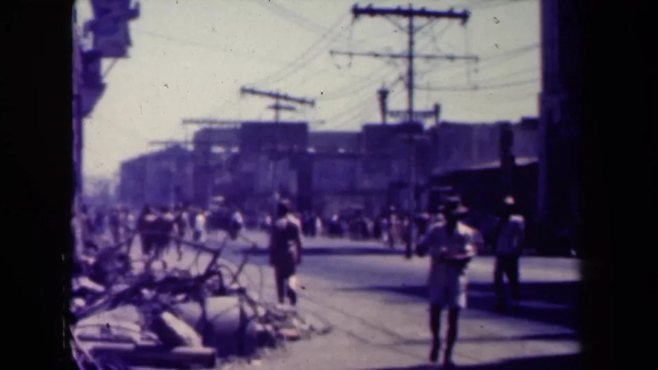 1944 Manila 8mm street frame showing pedestrians near rubble and overhead wires, with a figure standing at the right edge.