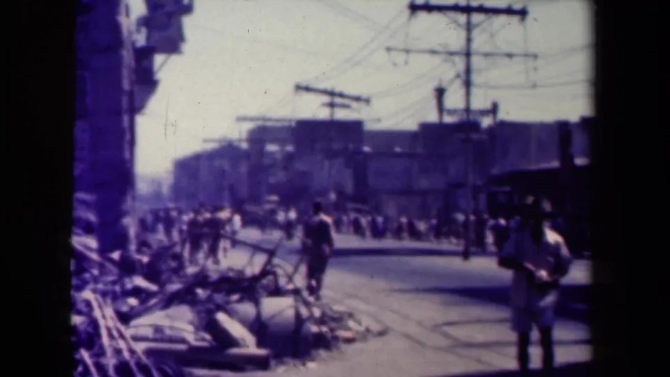 1944 Manila street footage frame with people walking past debris under overhead wires and damaged-looking urban structures.