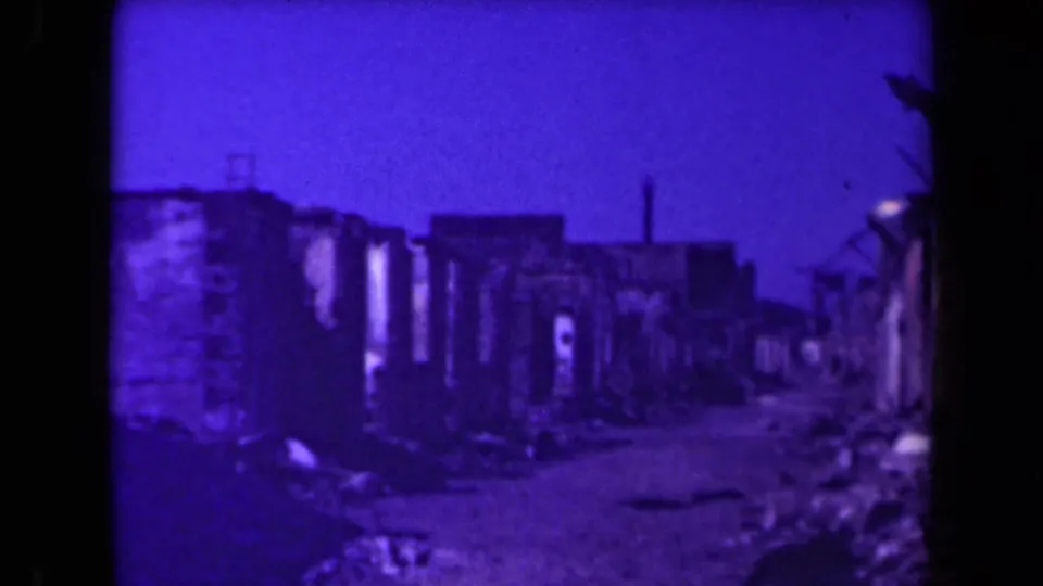 Purple-cast 8mm frame of a Manila ruin-lined road, with roofless walls, scattered debris, and a darkened street edge.