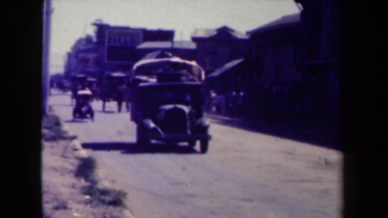 Vintage car approaches on a sunlit street with pedestrians and storefronts in the background.