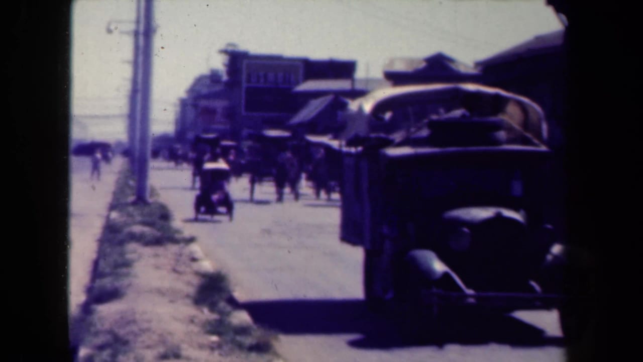 Car passes close to the camera as pedestrians move along a commercial street.
