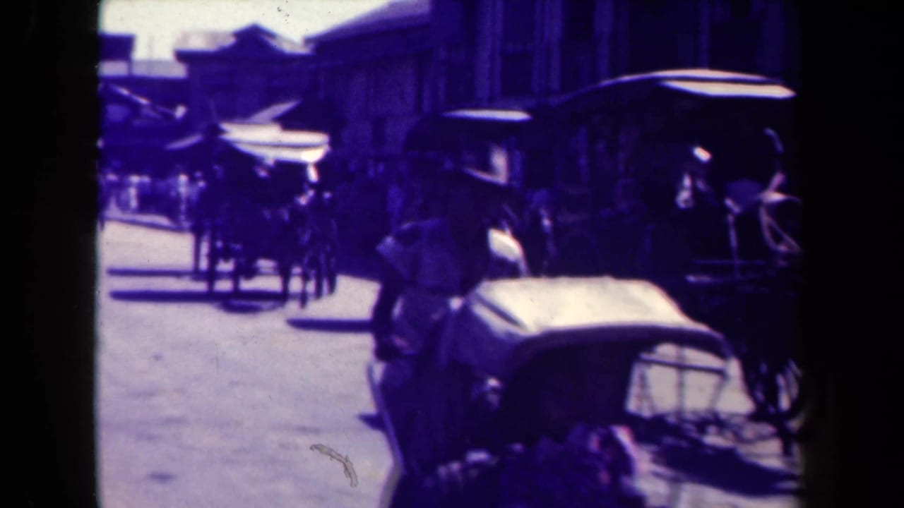 Automobile in the foreground with horse-drawn carriages farther along the street.