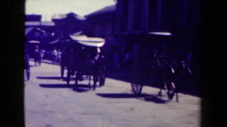 Horse-drawn carriage travels along a Manila street lined with awnings, pedestrians, and shadowed storefronts.