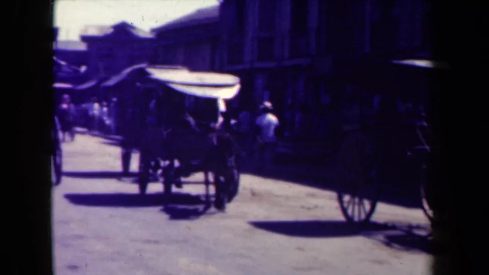 Horse-drawn carriage moves past the camera on a 1944 Manila street, with pedestrians and storefront shadows in the background.