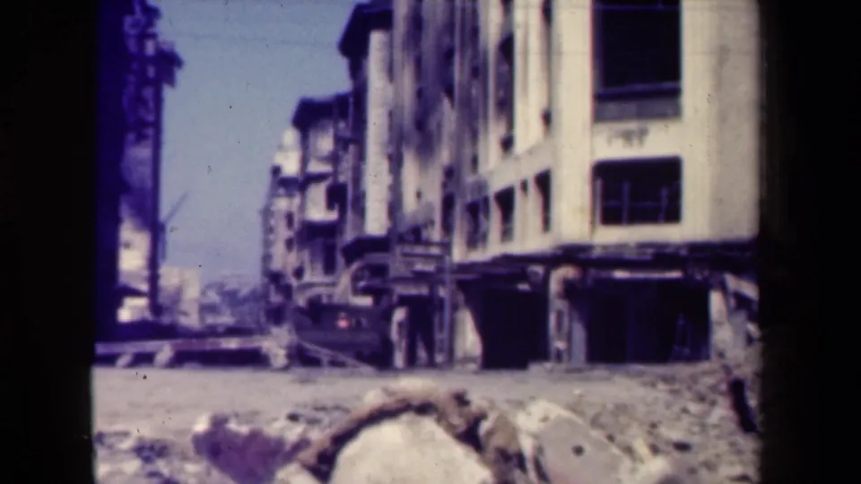 Dark truck beside a damaged white building in 1944 Manila, with rubble, open windows, and a utility pole in view.