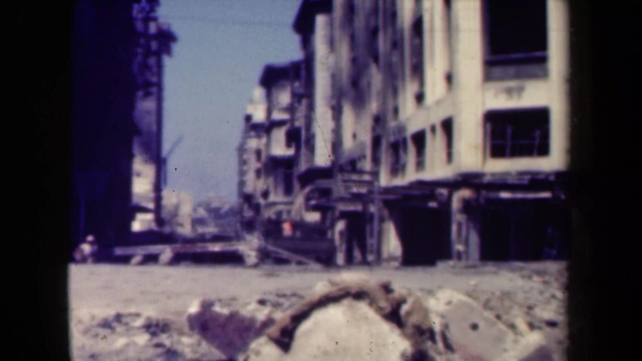 Damaged city buildings and rubble along a street with a few distant pedestrians.