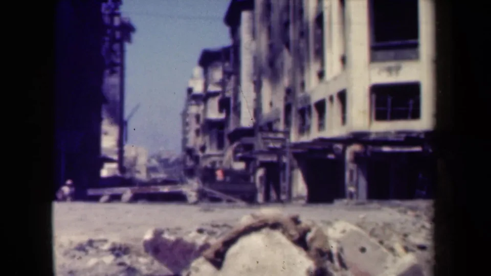 1940s Manila street view with damaged multi-story buildings, debris in the foreground, and distant pedestrians near the sidewalk.