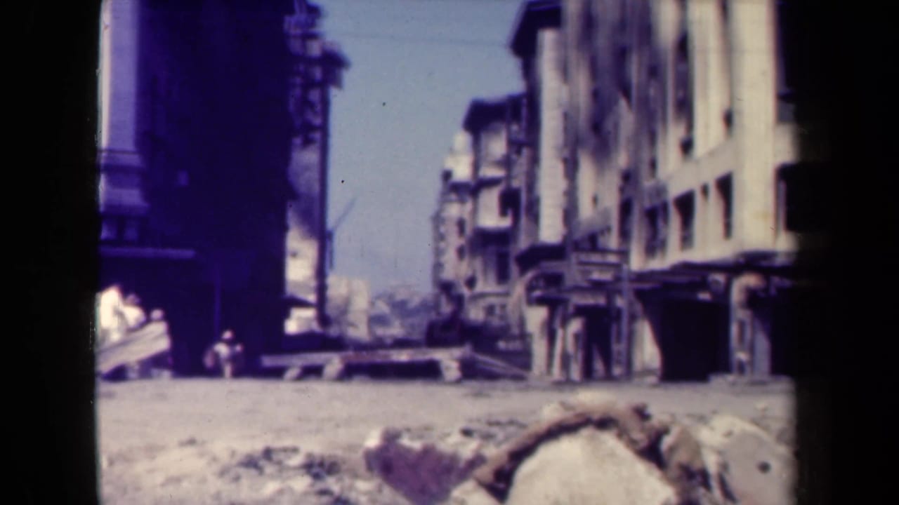 Street corner with damaged buildings, rubble, and pedestrians near the sidewalk.