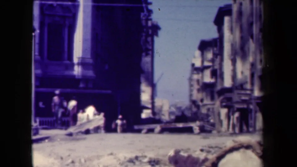 1940s Manila urban block showing broken facades, debris across the street, and civilians gathered or walking near a sidewalk.