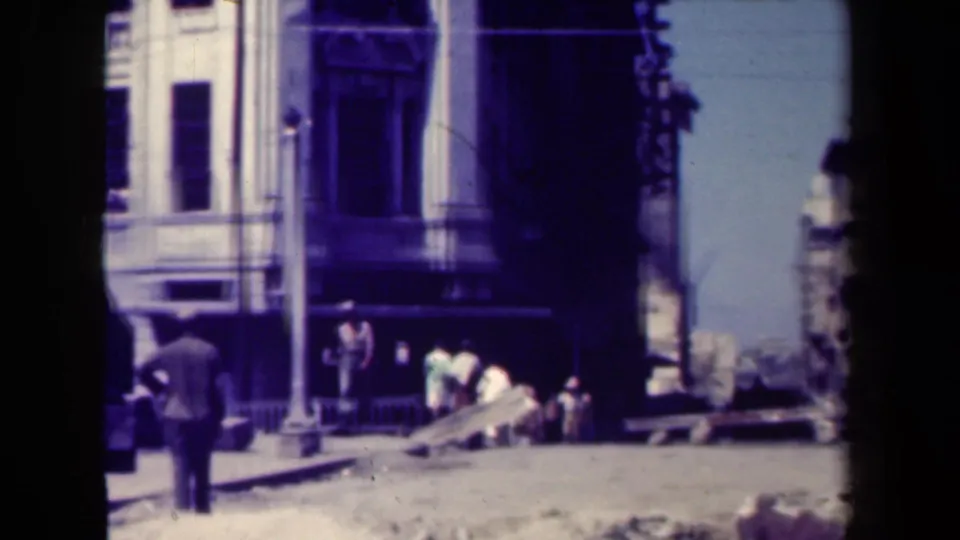 Street-level 1940s Manila view with a man seen from behind, damaged building fronts, rubble, and civilians near the sidewalk.