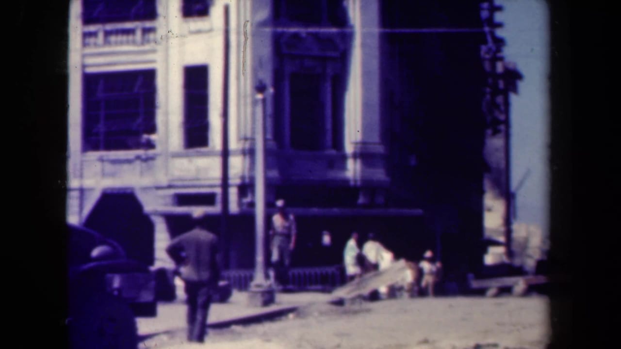 Man walking on a sunlit street near pedestrians, older cars, and multi-story building facades.