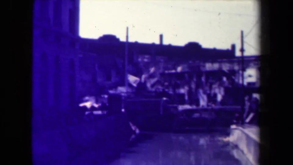 1944 Manila street view with shadowed storefronts, vertical poles, a dark vehicle-like shape, and distant urban structures in soft 8mm focus.