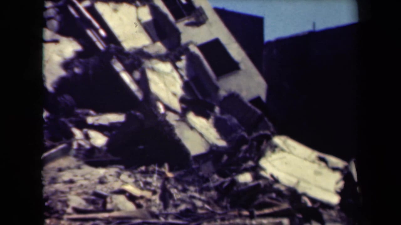 Collapsed building section with tilted wall, broken openings, and rubble below.