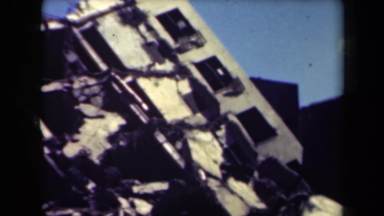 Damaged pale building with broken openings, exposed structure, and blue sky behind it.